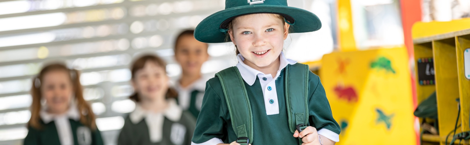 Young student in Holy Cross school uniform with backpack, standing in a bright classroom with other students in the background.